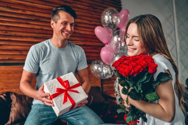 Sevgililer Günü 'nü kutluyoruz. Bir erkek bir kadına hediye ve kırmızı güller verir. Bir çift kalp şeklinde konfetiyle yatakta oturuyor..