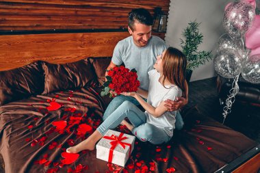 Sevgililer Günü 'nü kutluyoruz. Bir erkek bir kadına hediye ve kırmızı güller verir. Bir çift kalp şeklinde konfetiyle yatakta oturuyor..