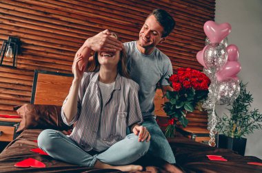 Yatak odasında güzel genç bir çift var. Sevgililer Günü 'nü kutluyoruz. Kırmızı gülleri olan bir adam bir kadının gözlerine elini kapatır ve sürpriz yapar..