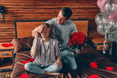 Yatak odasında güzel genç bir çift var. Sevgililer Günü 'nü kutluyoruz. Kırmızı gülleri olan bir adam bir kadının gözlerine elini kapatır ve sürpriz yapar..