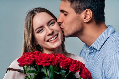 Gri arka planda izole edilmiş güzel romantik bir çift. Kırmızı güller tutan çekici genç bir kadın ve tişörtlü yakışıklı bir adam aşıklar. Sevgililer Günün kutlu olsun.!