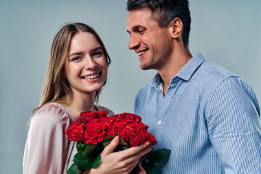 Gri arka planda izole edilmiş güzel romantik bir çift. Kırmızı güller tutan çekici genç bir kadın ve tişörtlü yakışıklı bir adam aşıklar. Sevgililer Günün kutlu olsun.!