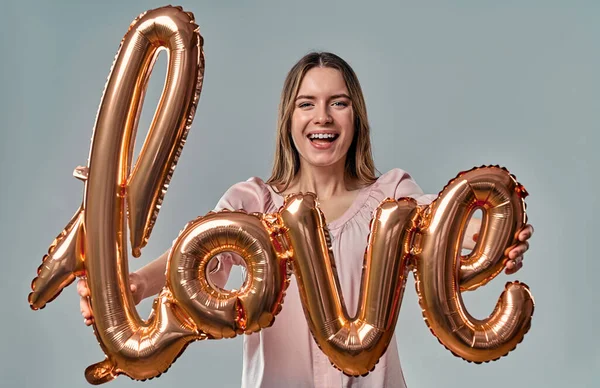 Attractive young woman in blouse is standing with air balloon labeled love in hands  on grey background.