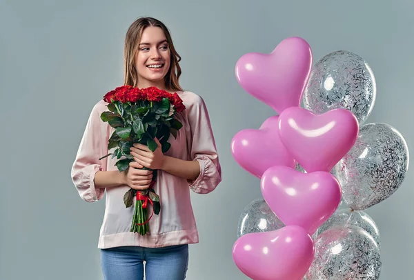 Portrait of attractive young woman in blouse is standing on grey background with red roses in hands and balloons.