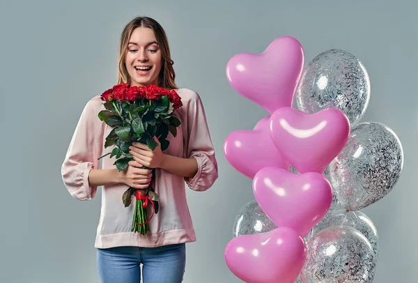Portrait of attractive young woman in blouse is standing on grey background with red roses in hands and balloons.