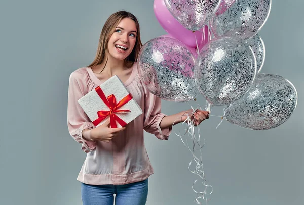 Portrait of attractive young woman in blouse is standing on grey background with gift box in hands and balloons.