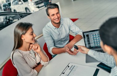 Genç çift araba alırken gülümsüyor, bir otomobil fuarının satış elemanı anahtar veriyor..