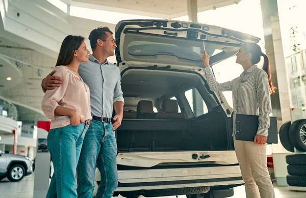 Buying their first car together. Young car saleswoman standing at the dealership telling about the features of the car to the customers.