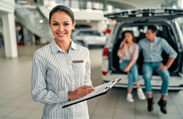 Young saleswoman consultant at a car dealership. In the background, the couple is sitting in the open trunk of their new car.