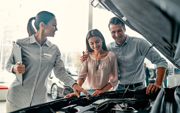 Buying their first car together. Young car saleswoman standing at the dealership telling about the features of the car under the hood to the customers.