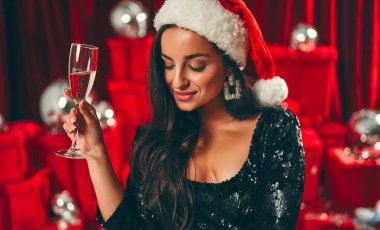 Mutlu yıllar sana! Noel Baba şapkalı, bir bardak şampanyayla güzel, seksi esmer kadın. Yeni yıl partisi. Noel arifesi. Kırmızı hediye kutuları ve disko toplarının arka planında.