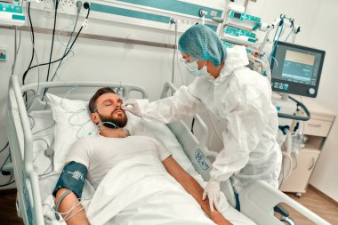 Koruyucu giysili, gözlüklü ve maskeli bir doktor yoğun bakım ünitesinde Coronavirus hastalığı olan bir hastanın durumunu gözlemliyor. Modern bir klinikte tedavi.