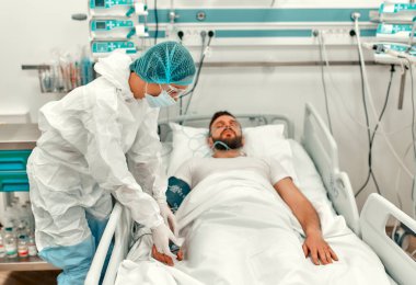 Koruyucu giysili, gözlüklü ve maskeli bir doktor yoğun bakım ünitesinde Coronavirus hastalığı olan bir hastanın durumunu gözlemliyor. Modern bir klinikte salgın sırasında tedavi.
