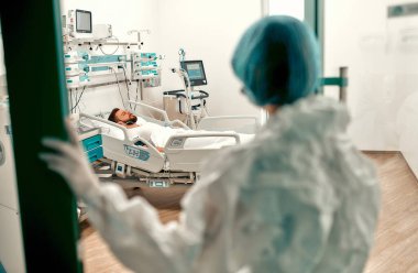 Koruyucu kıyafetli bir doktor, modern bir hastanede yoğun bakım ünitesinde bulunan Coronavirus hastalığı olan bir adamı kontrol ediyor..
