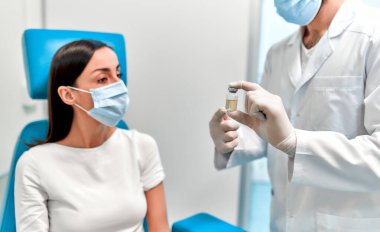 Aşı, aşı kampanyası, hastalık önleme konsepti. Tıbbi maske takan bir kadın, Covid-19 aşısını bir doktorun ofisinde alır. Hemşire hastaya aşıdan bahseder..