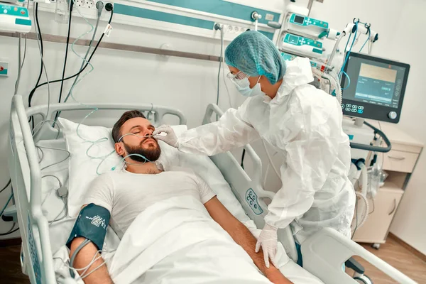 Koruyucu giysili, gözlüklü ve maskeli bir doktor yoğun bakım ünitesinde Coronavirus hastalığı olan bir hastanın durumunu gözlemliyor. Modern bir klinikte tedavi.