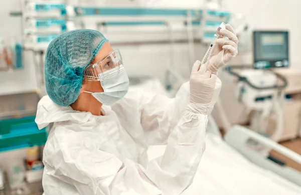 A doctor in a protective suit holds a syringe and an ampoule with the covid-19 coronavirus vaccine while standing in an intensive care unit in a modern hospital.