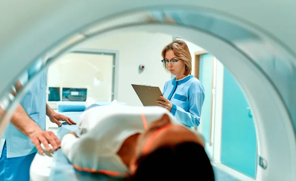 A patient lying on CT or MRI, the bed moves inside the machine, scanning her body and brain under the supervision of a doctor and a radiologist. In a medical laboratory with high-tech equipment.