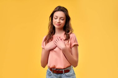 Genç, güzel, esmer, sıradan bir gömlek giyen, beyaz arka plan üzerinde duran, elleri göğsünde, gözleri kapalı ve yüzünde minnet dolu bir jest olan bir kadın. Sağlık kavramı.