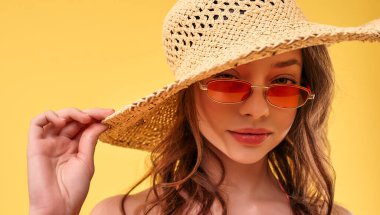 Mayo giymiş, sarı arka planda saman şapkalı ve güneş gözlüklü hoş seksi bir kadın. Seyahat, tatil, turizm konsepti.