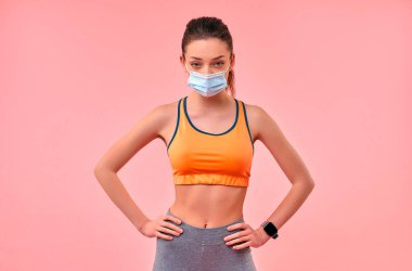 Pembe arka planda izole edilmiş koruyucu bir tıbbi maske takan spor giysili tatlı bir kız. Spor, yoga, karantina koşullarında aktif yaşam tarzı.
