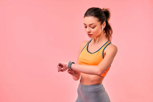 スポーツウェアのかわいいスポーティーな女の子は 時計の時間をチェックし ピンクの背景に隔離されたヘッドフォンで音楽を聴く スポーツ アクティブなライフスタイル 健康のための運動 — ストック写真