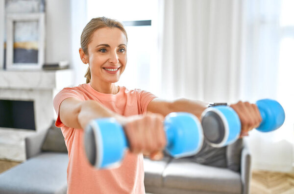 Sport, fitness and healthy lifestyle concept. Mature woman doing exercises with dumbbells at home. How to stay healthy in quarantine.