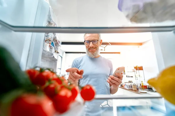 Elinde telefon olan olgun bir adam buzdolabını bir şeyler pişirmek ya da erzakları kontrol etmek için açtı. Buzdolabından bak. Sağlıklı gıda konsepti. Evde yemek sipariş etmek..