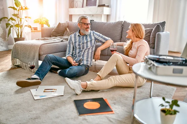 A mature couple are sitting on the floor near the sofa with glasses of wine, relaxing, enjoying life at home and listening to vinyl records on a music player. Romantic evening for a married couple.