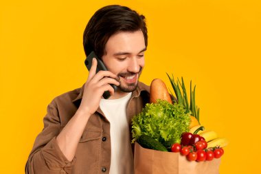 Elinde bir paket sebze ürünüyle telefonda konuşan sakallı genç adam turuncu arka planda izole edilmiş. İnternetten yemek siparişi vermek..