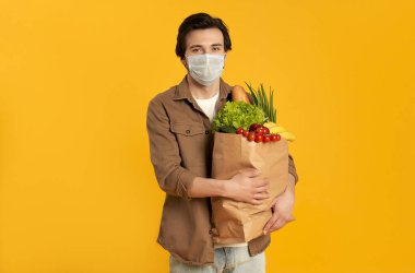 Koruyucu tıbbi maskeli genç sakallı bir adam. Elinde turuncu arka planda izole edilmiş sebze ürünleriyle dolu bir kese var..