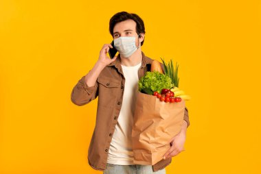 Koruyucu tıbbi maskeli genç sakallı adam. Elinde turuncu arka planda izole bir şekilde telefonla konuşan sebze ürünleri paketi var..