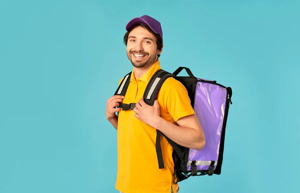 Portrait of a young courier, delivery man in uniform with a thermo backpack isolated on a blue background. Fast home delivery. Online order.