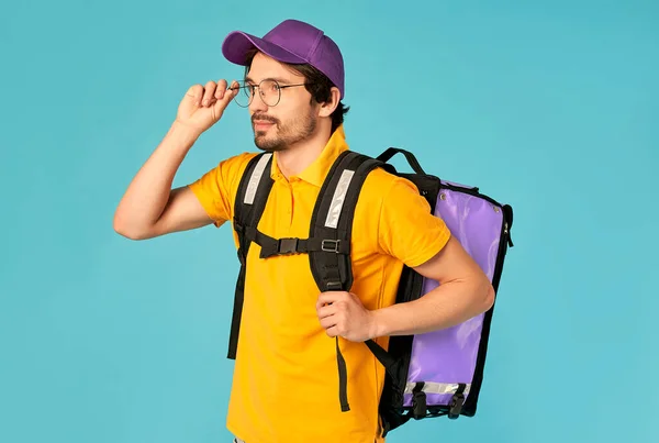 Portrait of a young courier, delivery man in uniform with a thermo backpack isolated on a blue background. Fast home delivery. Online order.