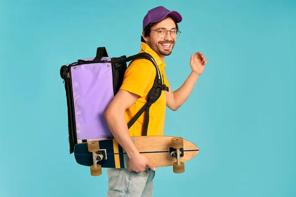 Young courier delivery man in uniform with thermo backpack and skate isolated on blue background. Fast express home delivery. Online order.
