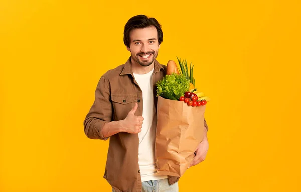Young bearded man with paper package of vegetables products shows thumbs up gesture isolated on orange background.