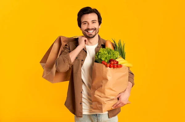 Young bearded man with paper bag of vegetables groceries and purchases isolated on orange background.