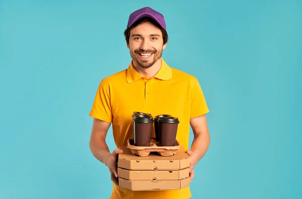 制服を着た若いひげを生やした宅配便の配達人は 青い背景に隔離されたピザとコーヒーの箱を持っています 宅配便 — ストック写真