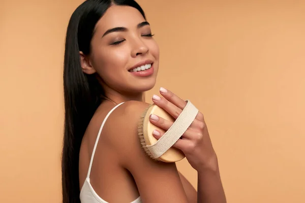 Young Asian woman in white lingerie and clean radiant skin makes anti-cellulite massage with a dry brush on a beige background. Body scrubbing, weight loss, body care, spa treatments.