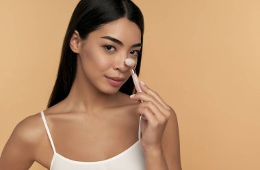 Temiz ciltli genç bir Asyalı kadın bej arka planda kuvars silindirli yüz masajı yapıyor. Spa bakımı, yüz bakımı, güzellik kozmetolojisi.