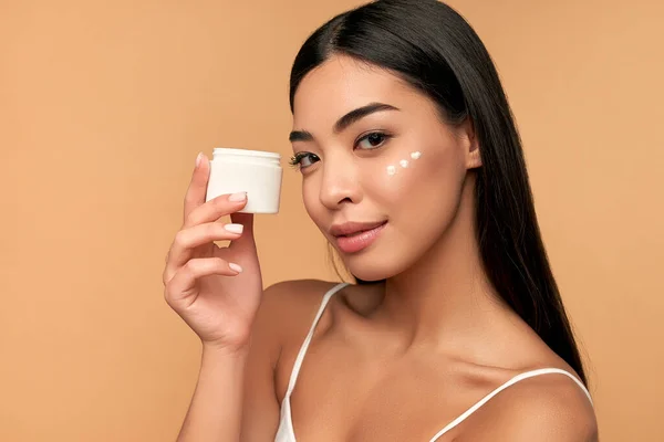 Young Asian woman with clean radiant skin uses a moisturizing face cream on a beige background. Spa care, facial skin care, beauty cosmetology.