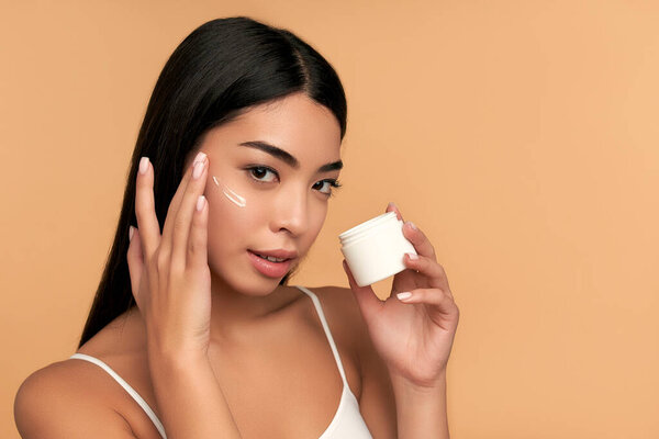 Молодая азиатка с чистой сияющей кожей использует увлажняющий крем для лица на бежевом фоне. СПА уход, уход за кожей лица, косметология красоты.