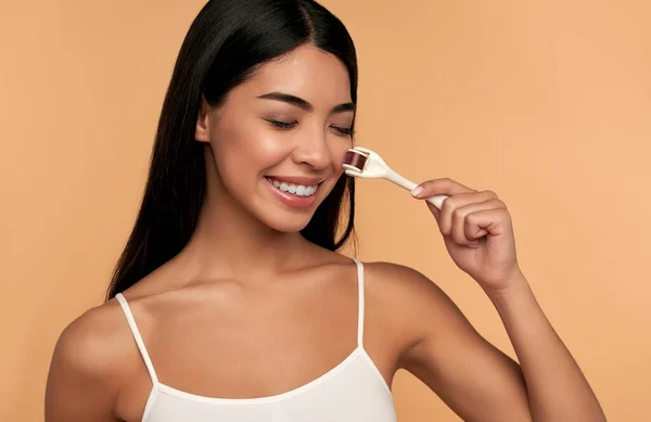 Mezoterapi prosedürleri için dermaroller kullanan Asyalı kadın yüzü, evde ve kuaförde cilt bakımı. Bej arka planda mikro kenarlı Mezoscooter.