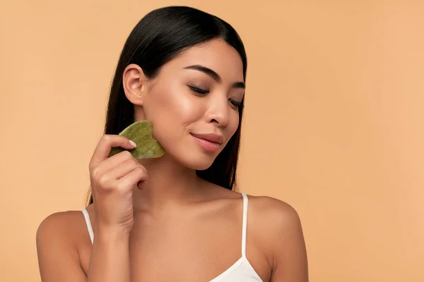 Gua Sha kuvars taşıyla masaj için güzel bir Asyalı kız. Bej arka plan izole edildi. Kozmetik doğal akım. Geleneksel tıp ve kozmetoloji kavramı.