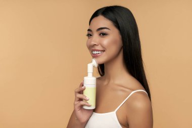 Parlak tenli genç Asyalı kadın yüzünü fırça ve köpükle temizliyor.