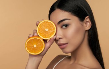 Bej arka planda izole edilmiş beyaz iç çamaşırlarıyla turuncu yarımlar tutan parlak ciltli Asyalı bir kızın portresi. C vitamini kozmetik konsepti