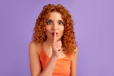 Kıvırcık saçlı, tatlı bir kız parmağını dudaklarına dayayıp usulca konuşuyor mor arka planda izole bir şekilde..