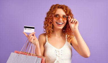 Kırmızı saçlı, kıvırcık saçlı ve elinde mor arka planda izole edilmiş bir kredi kartı olan hoş bir kız. İnternetten alışveriş. Satılık. Alışveriş konsepti.