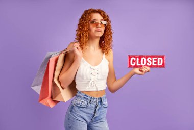 Kıvırcık saçlı, elinde mor arka planda kapalı olduğunu söyleyen bir tabela tutan sevimli, üzgün, kızıl saçlı kız. Alışveriş konsepti, alışveriş, satış..