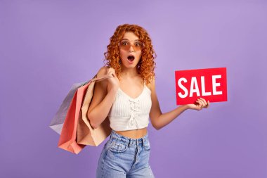 Kırmızı saçlı, kıvırcık saçlı, elinde mor arka planda satıştan izole edilmiş bir tabela tutan hoş bir kız. İnternetten alışveriş. Satılık. Alışveriş konsepti.